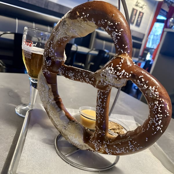 A large, salted pretzel on a stand with dipping sauces and a glass of beer at Outpost 611 Eatery.